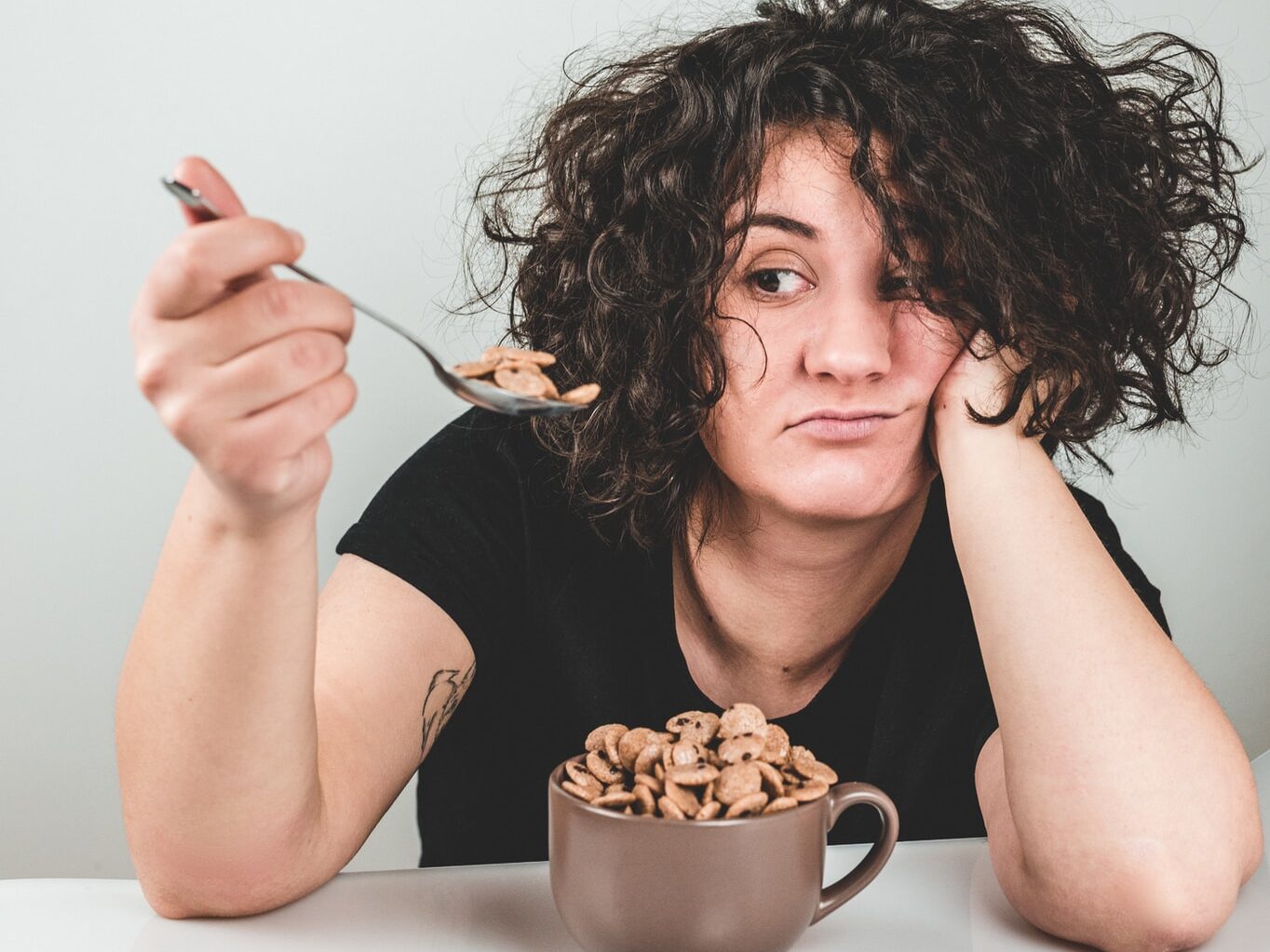 woman with messy hair wearing black crew-neck t-shirt holding spoon with cereals on top