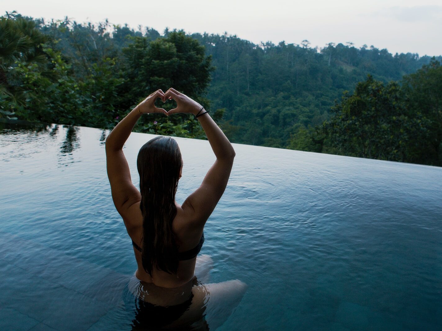 - ČasProŽeny.cz woman in infinity pool making heart hand gesture facing green leafed trees