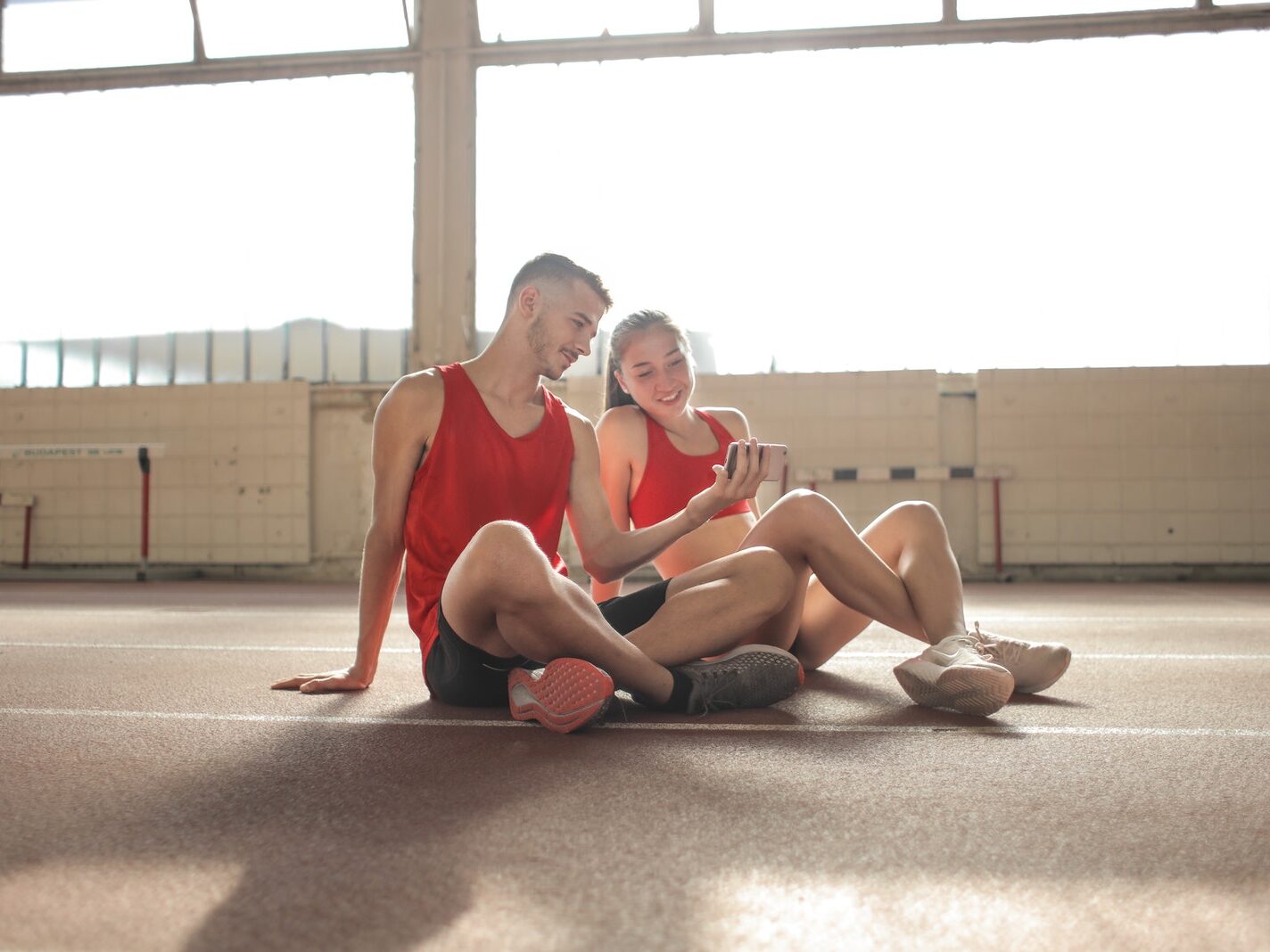- ČasProŽeny.cz Happy young sportsmen sitting with crossed legs on floor and surfing smartphone together at sports hall