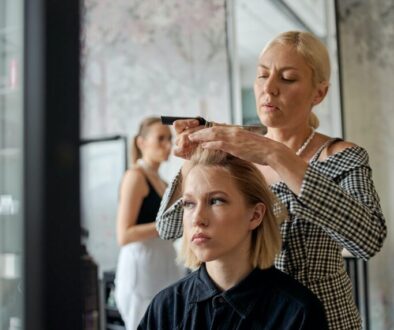 Adult female hairstylist straightening hair of dreamy client with iron reflecting in mirror in beauty salon
