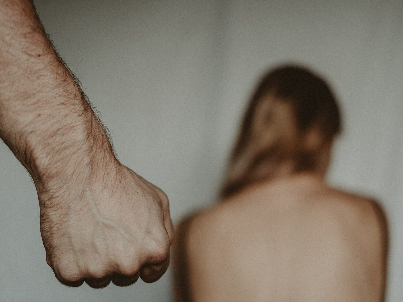 Aggressive abusive anonymous man clenching fist while insulting unrecognizable vulnerable female sitting on blurred background in light room during violence