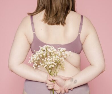 Back view of anonymous plump female in bra and panties demonstrating curves of body standing with bunch of fresh flowers against pink background