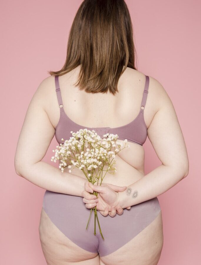 Back view of anonymous plump female in bra and panties demonstrating curves of body standing with bunch of fresh flowers against pink background