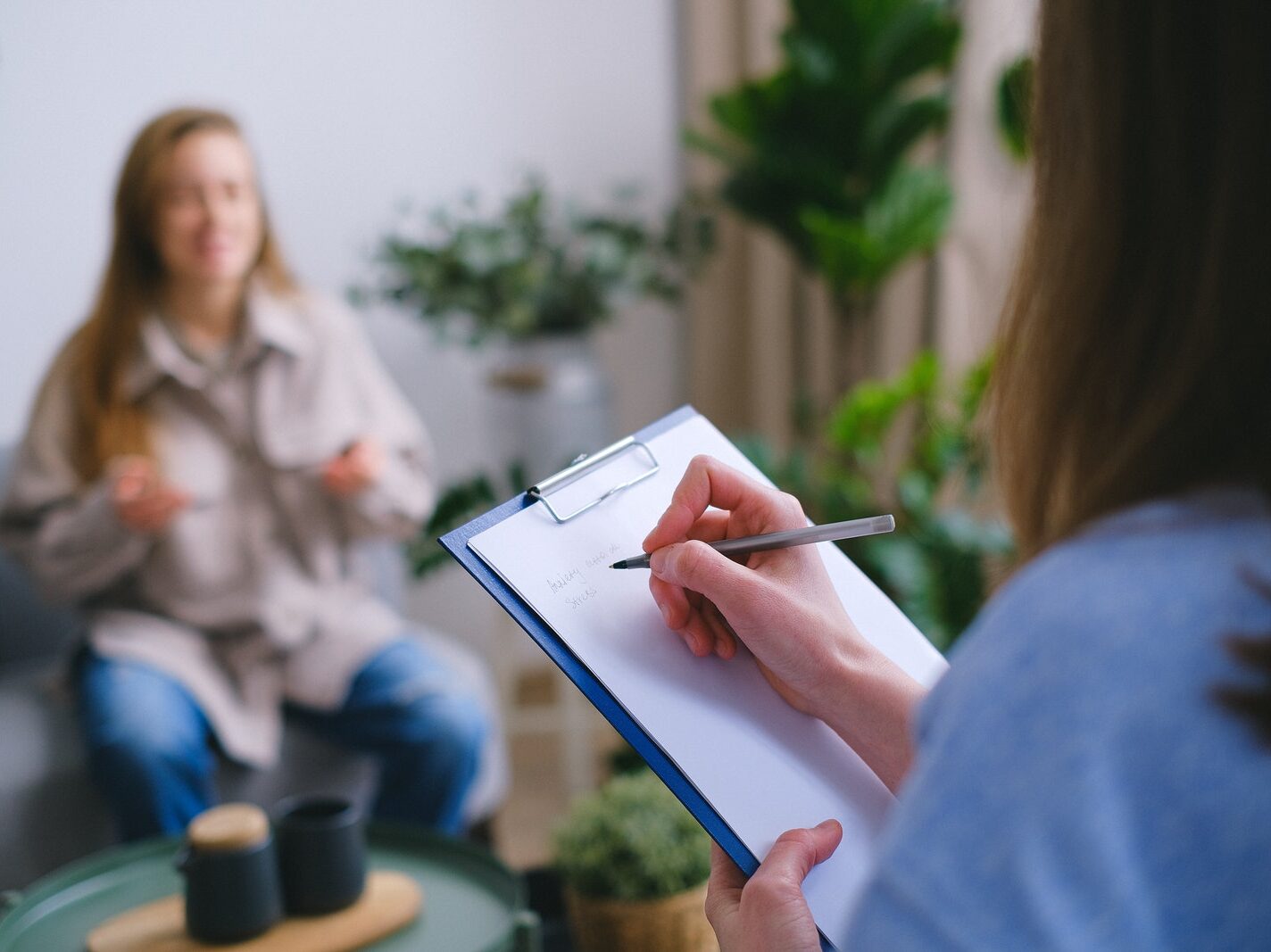 Unrecognizable professional female psychologist writing on clipboard while sitting against client on blurred background during psychotherapy session in light office