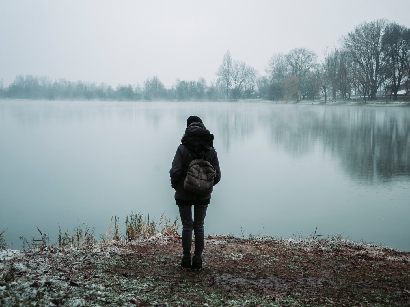 person wearing black hooded jacket standing and facing on body of water near trees in foggy day