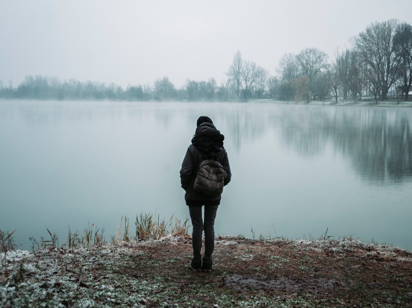 person wearing black hooded jacket standing and facing on body of water near trees in foggy day