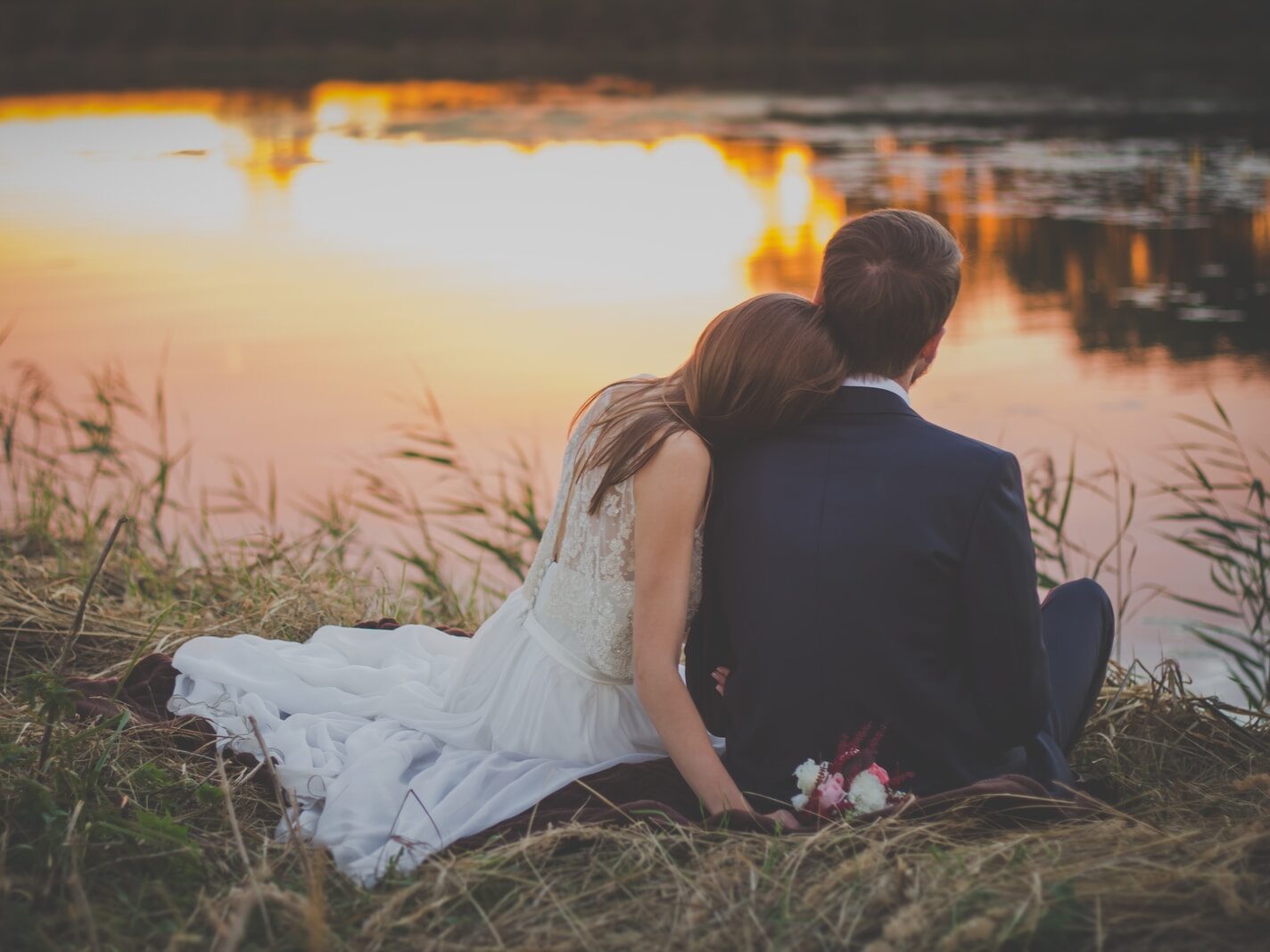 sitting woman leaning on man's shoulder facing lake during golden hour