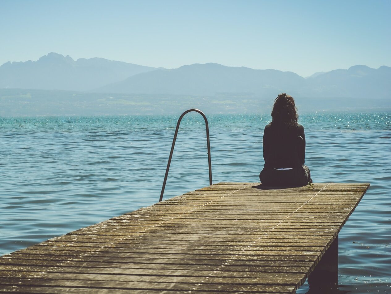 a woman sits on the end of a dock during daytime staring across a lake