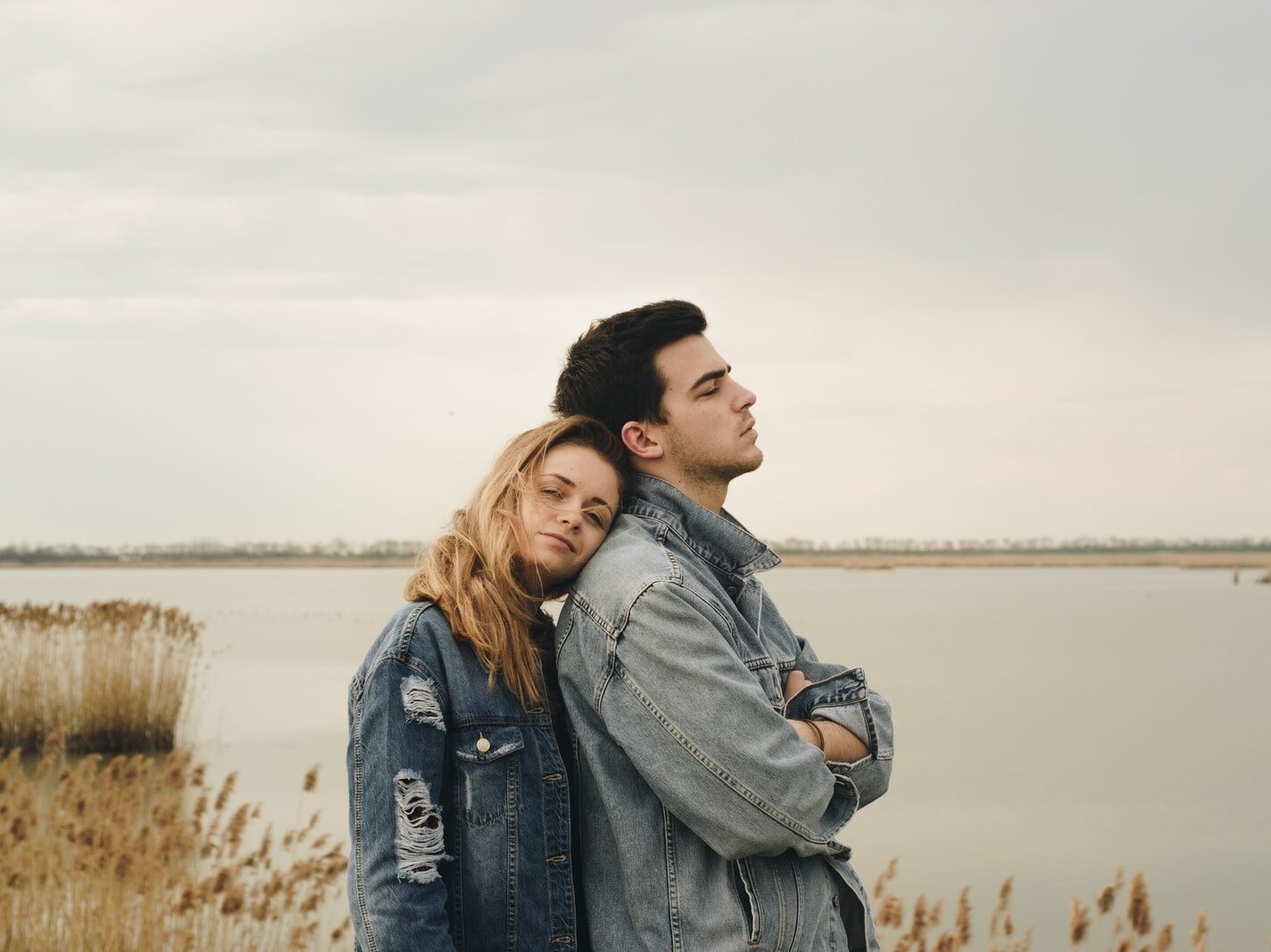 woman in blue denim jacket leaning on man's shoulder near body of water
