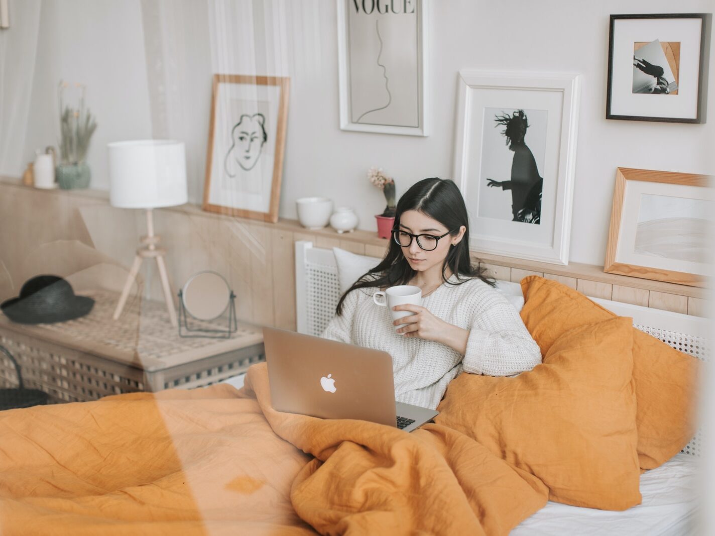 Focused young brunette in eyeglasses with cup of hot drink lying in comfortable bed and working on laptop in morning