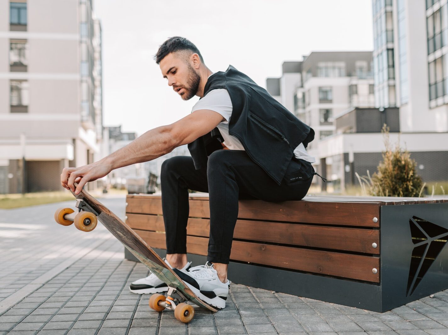 A Man in Black Pants Sitting on a Wooden Bench while Stepping on His Skateboard