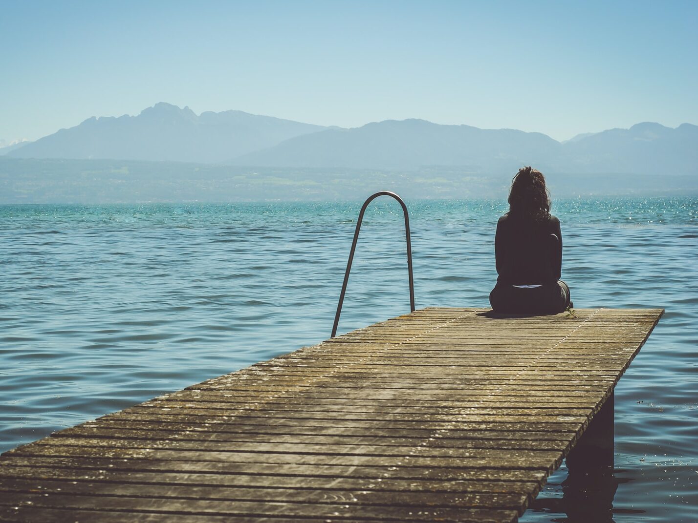 a woman sits on the end of a dock during daytime staring across a lake