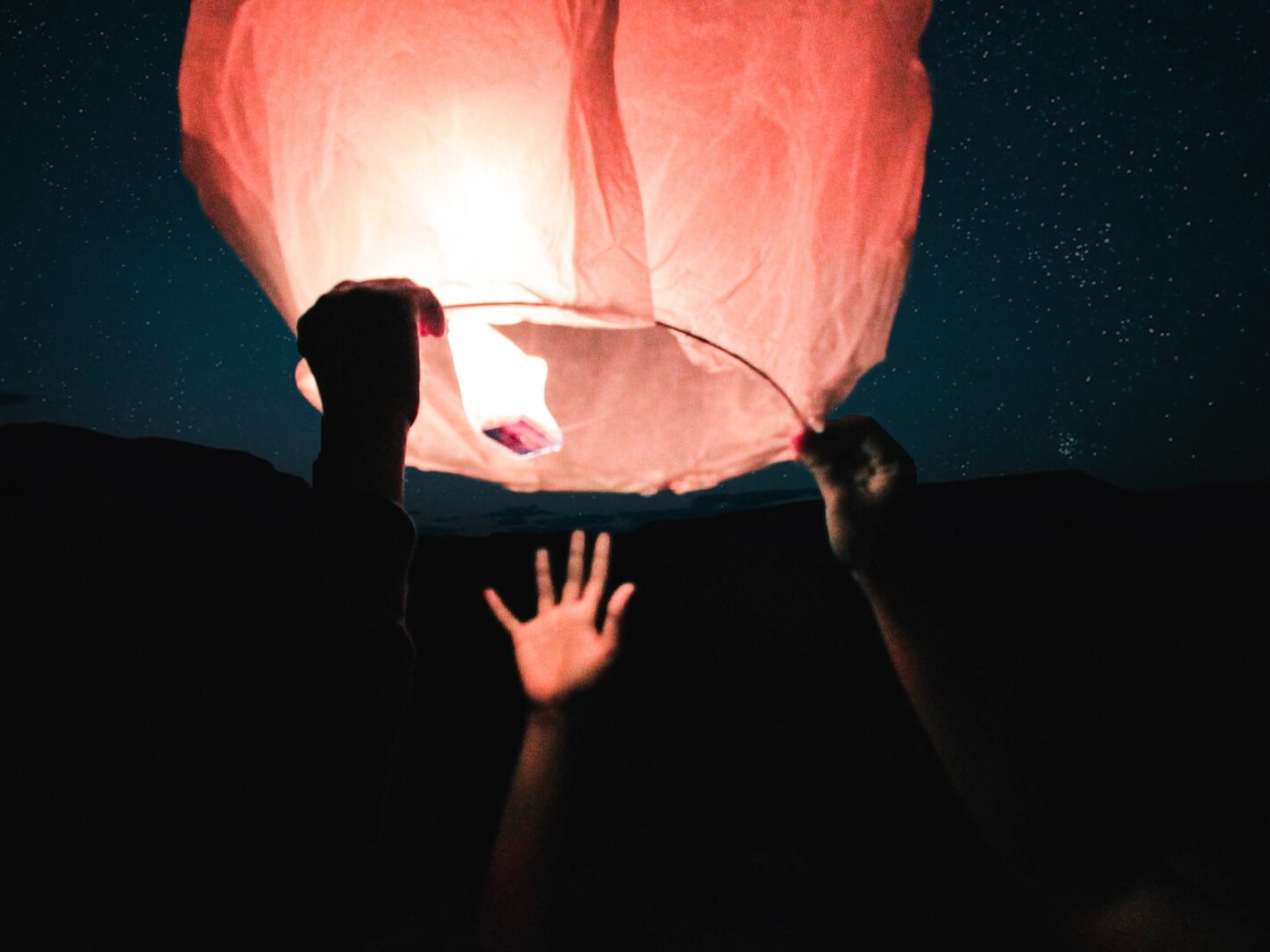 three human hand about to catch and hold sky lantern at night time