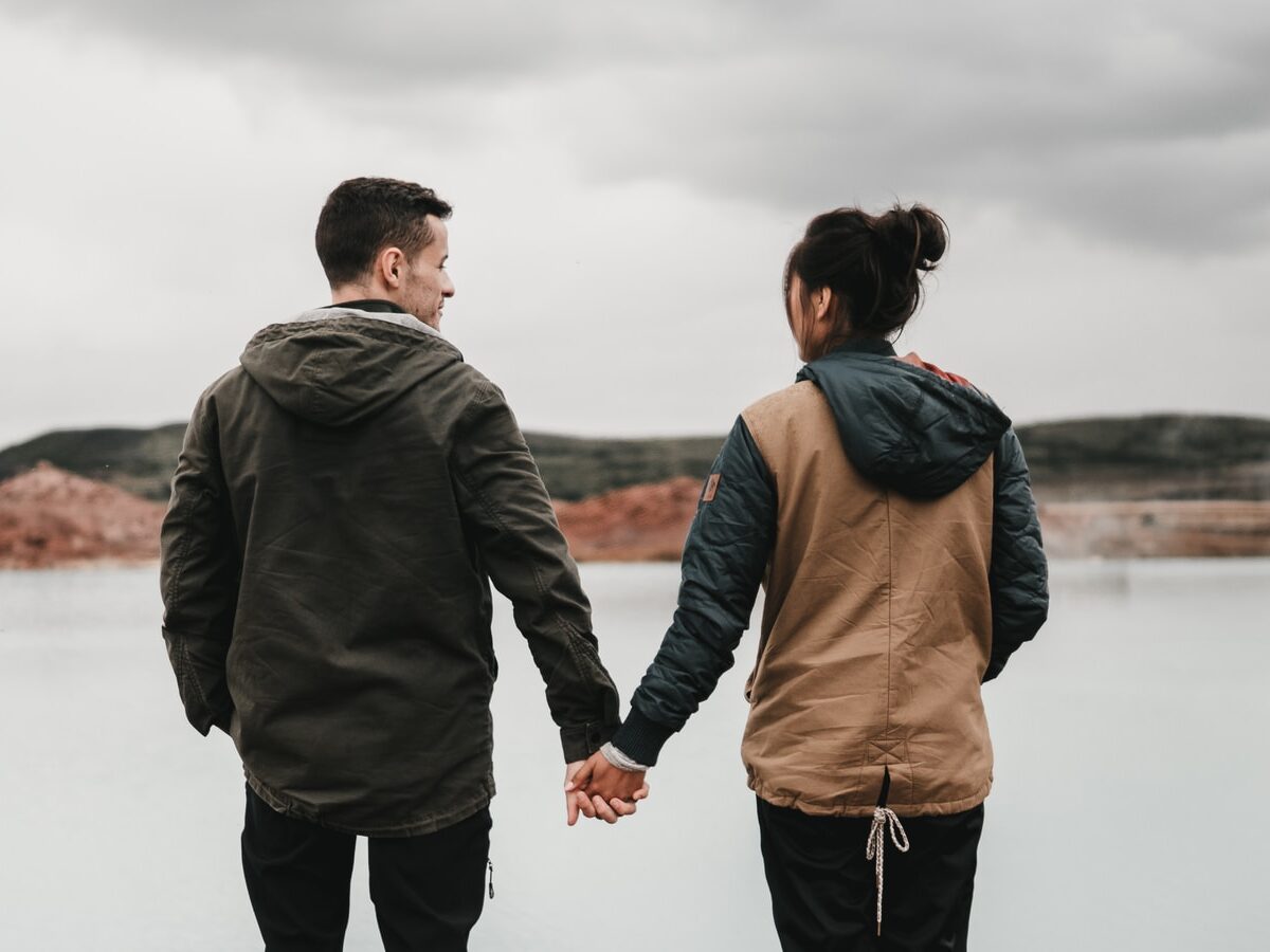 couple holding hand front of body of calm water with mountain distance