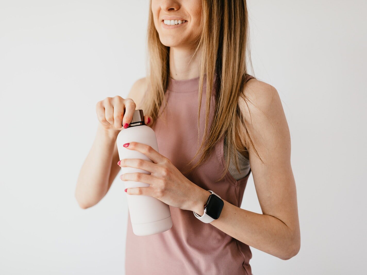 Crop faceless young female in casual outfit wearing smart watch opening cosmetic bottle while standing against white background