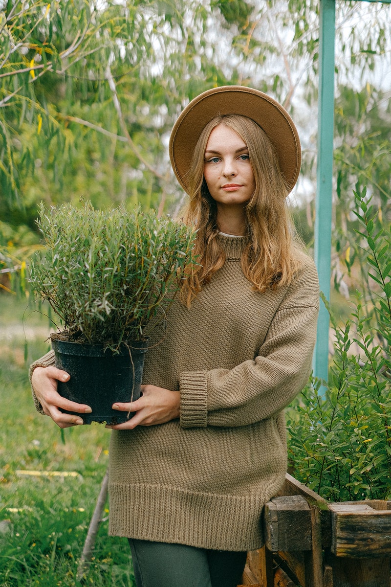 Young positive female gardener wearing brown sweater and hat holding lush potted plant while standing in verdant botanical garden and looking at camera