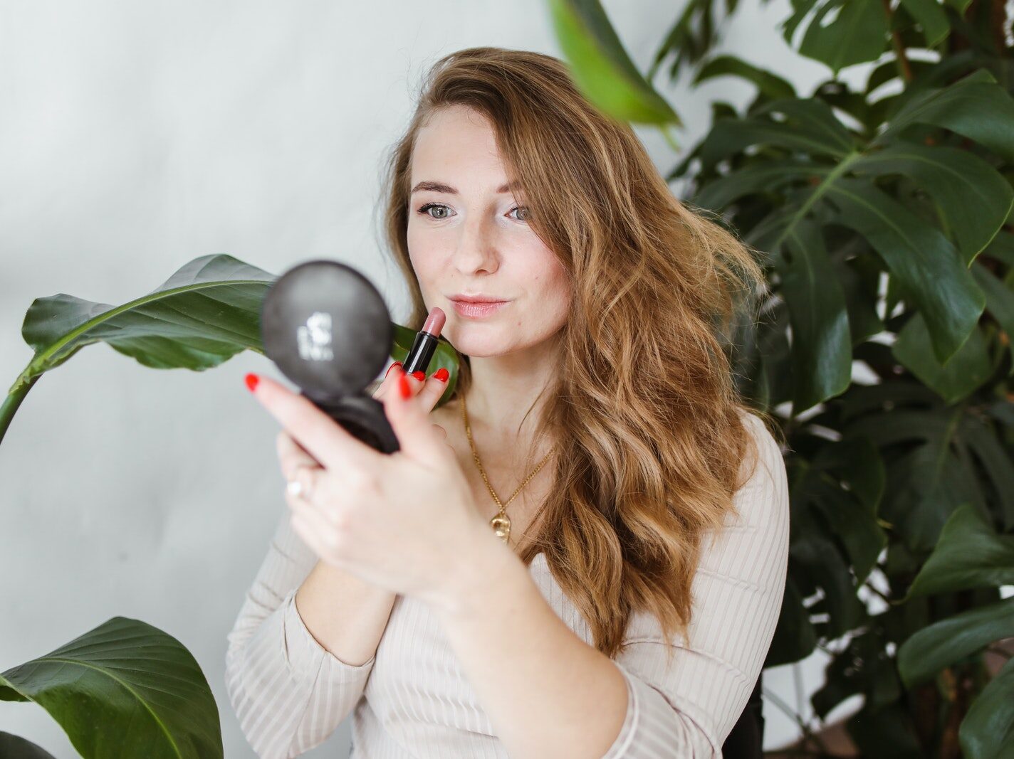 Photo of a Woman with Blond Hair Putting on Pink Lipstick Near Green Plants