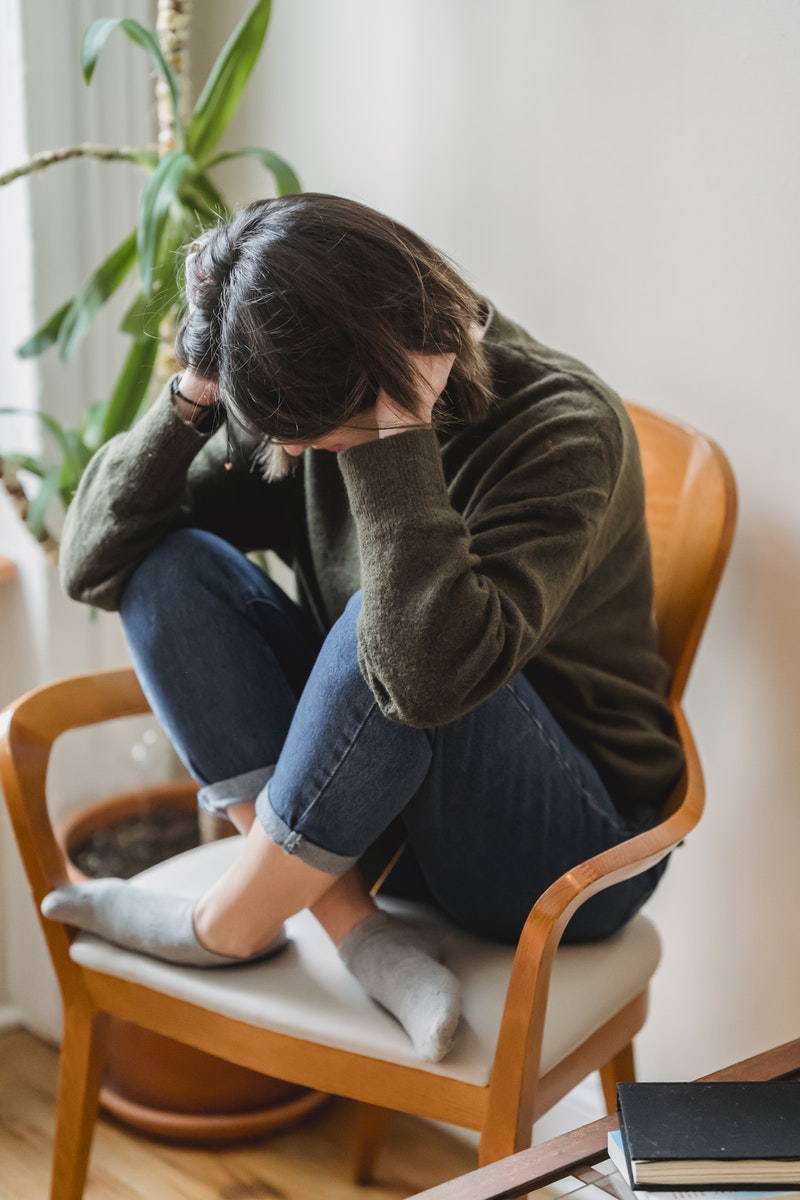 Full body of unrecognizable young worried female in casual clothes sitting on chair with crossed legs and covering ears with hands at home