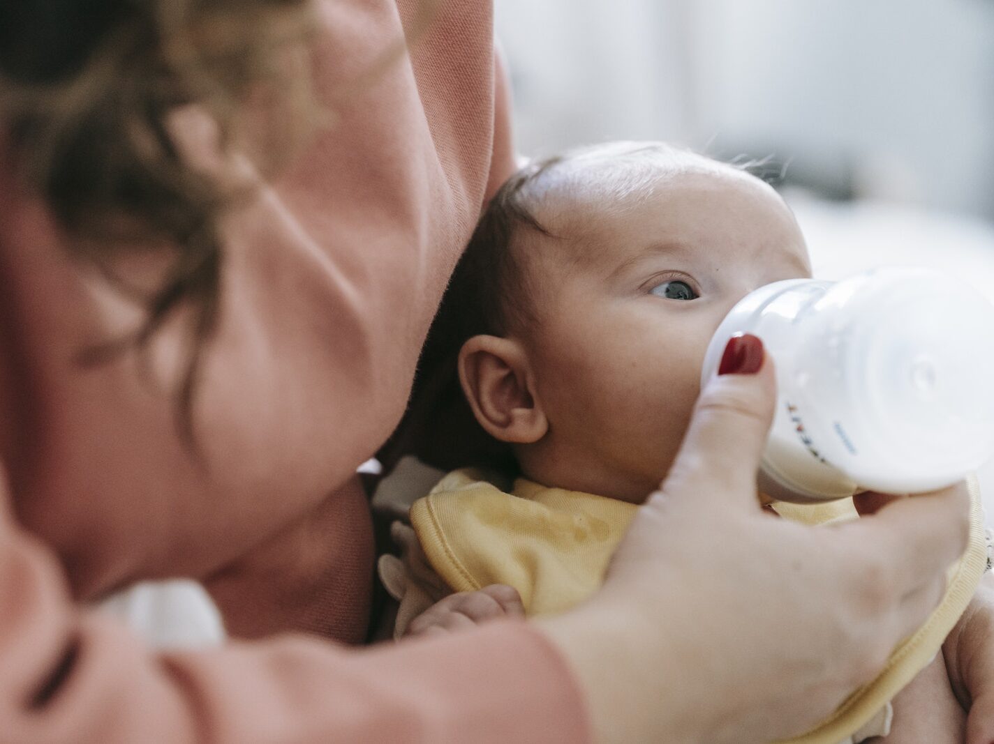 Crop faceless mother in casual clothes embracing and feeding cute newborn baby with milk from bottle in daylight at home