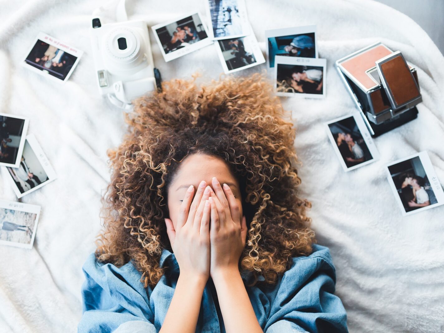 woman lying on bed covering her face surrounded by photos and white camera