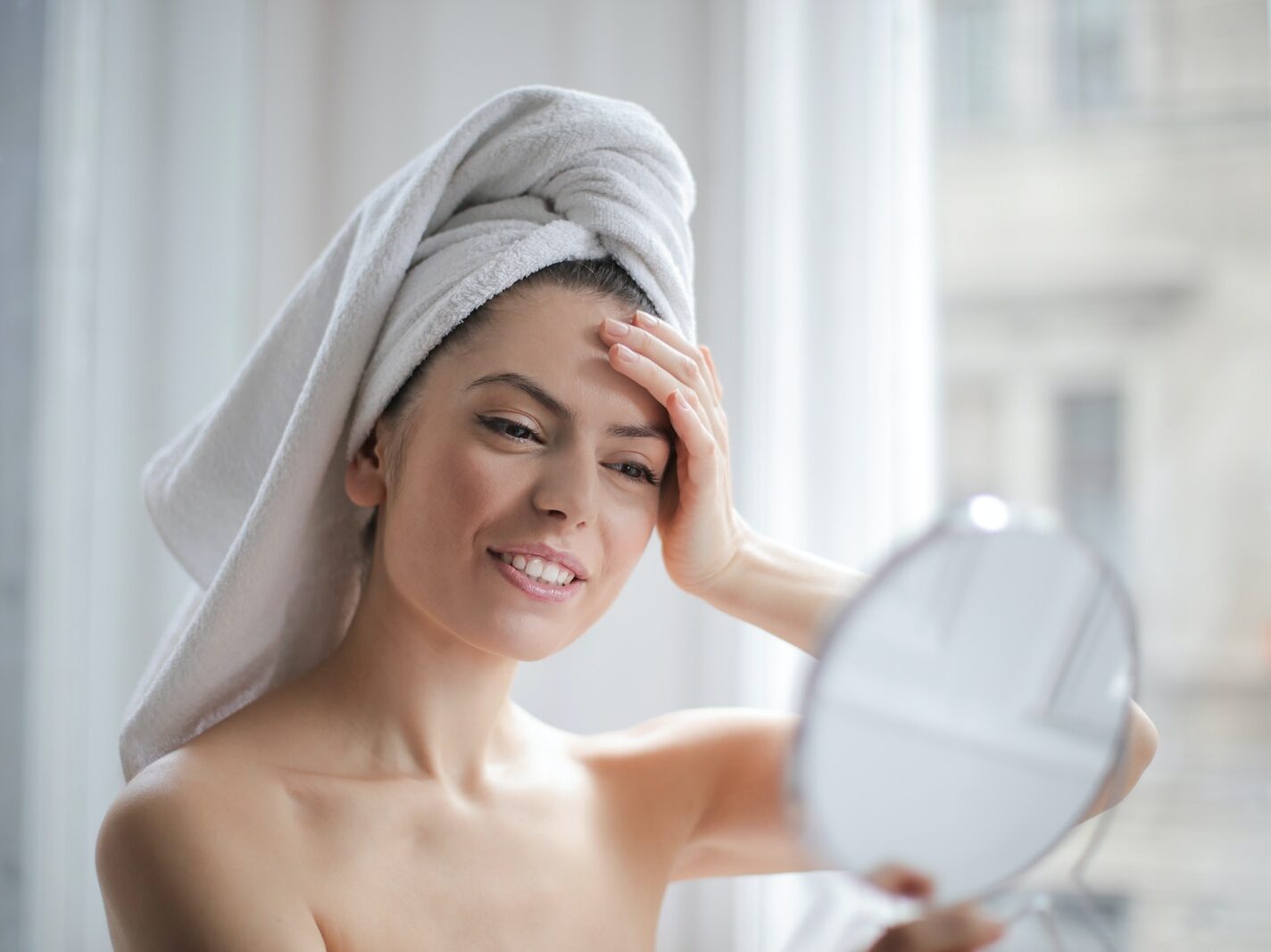 Selective Focus Portrait Photo of Smiling Woman With a Towel on Head Looking in the Mirror