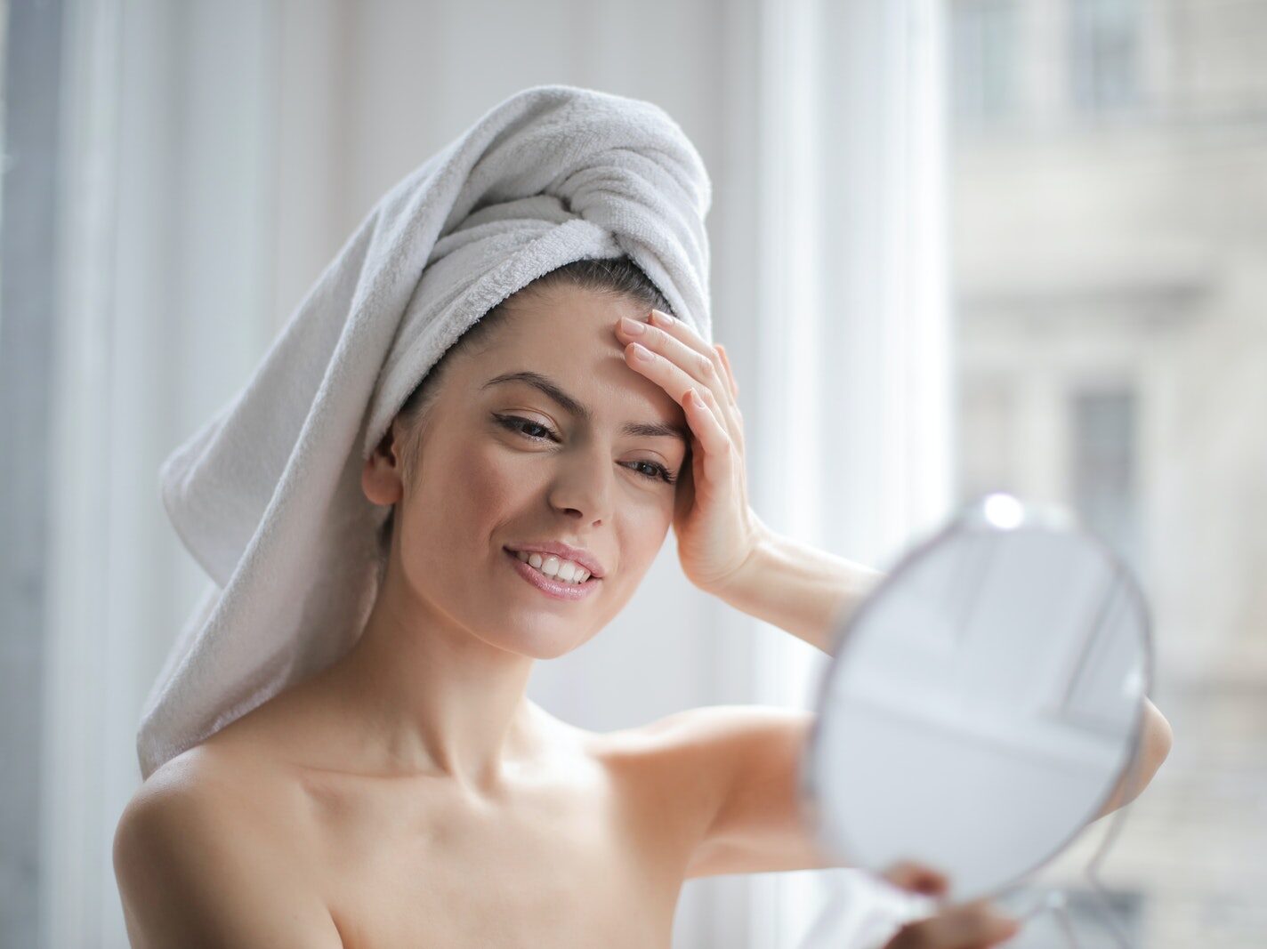 Selective Focus Portrait Photo of Smiling Woman With a Towel on Head Looking in the Mirror