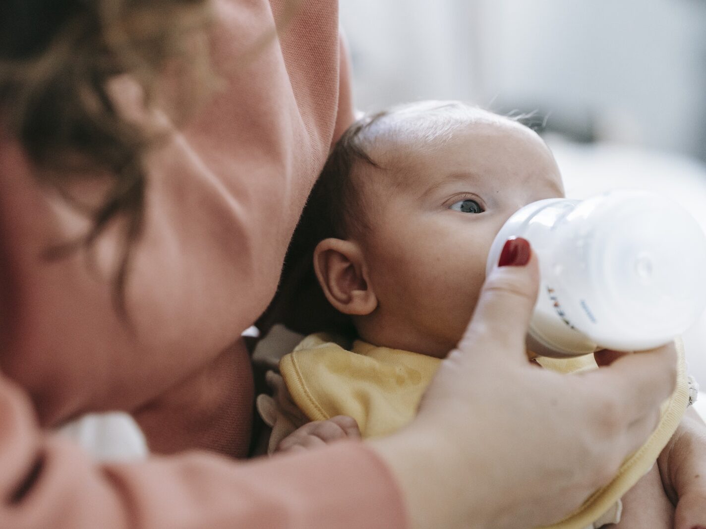 Crop faceless mother in casual clothes embracing and feeding cute newborn baby with milk from bottle in daylight at home