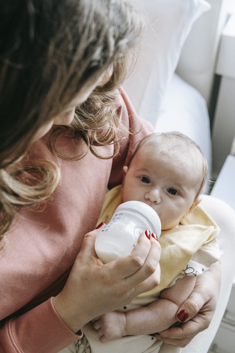 High angle of crop unrecognizable young mother in casual clothes sitting on bed and feeding adorable baby with milk from bottle in sunny morning