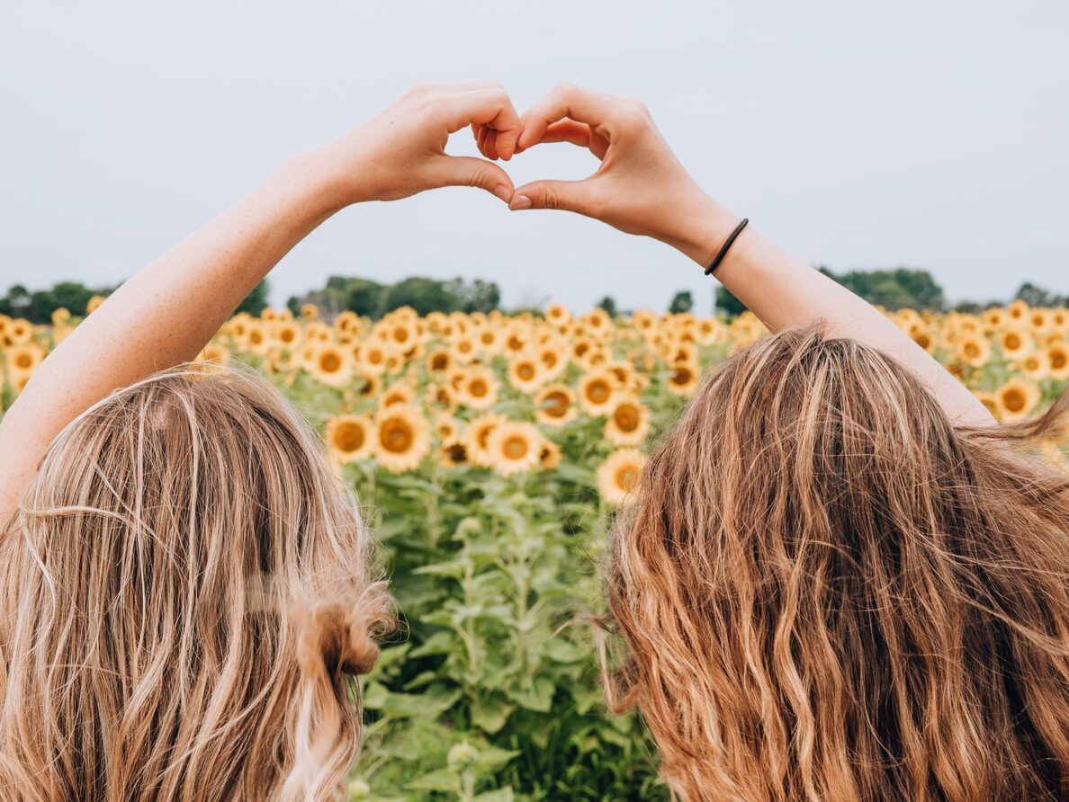 two women forming heart-shape using hands fronting sunflower field during daytime