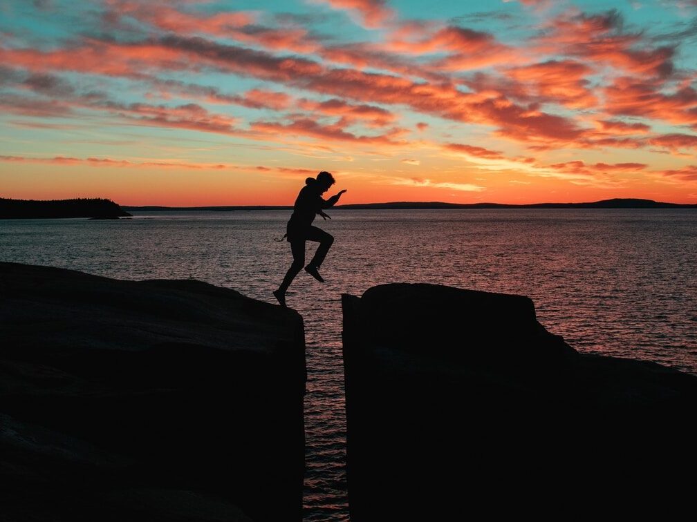 silhouette photo of a man jumps on to cliff near sea during sunset