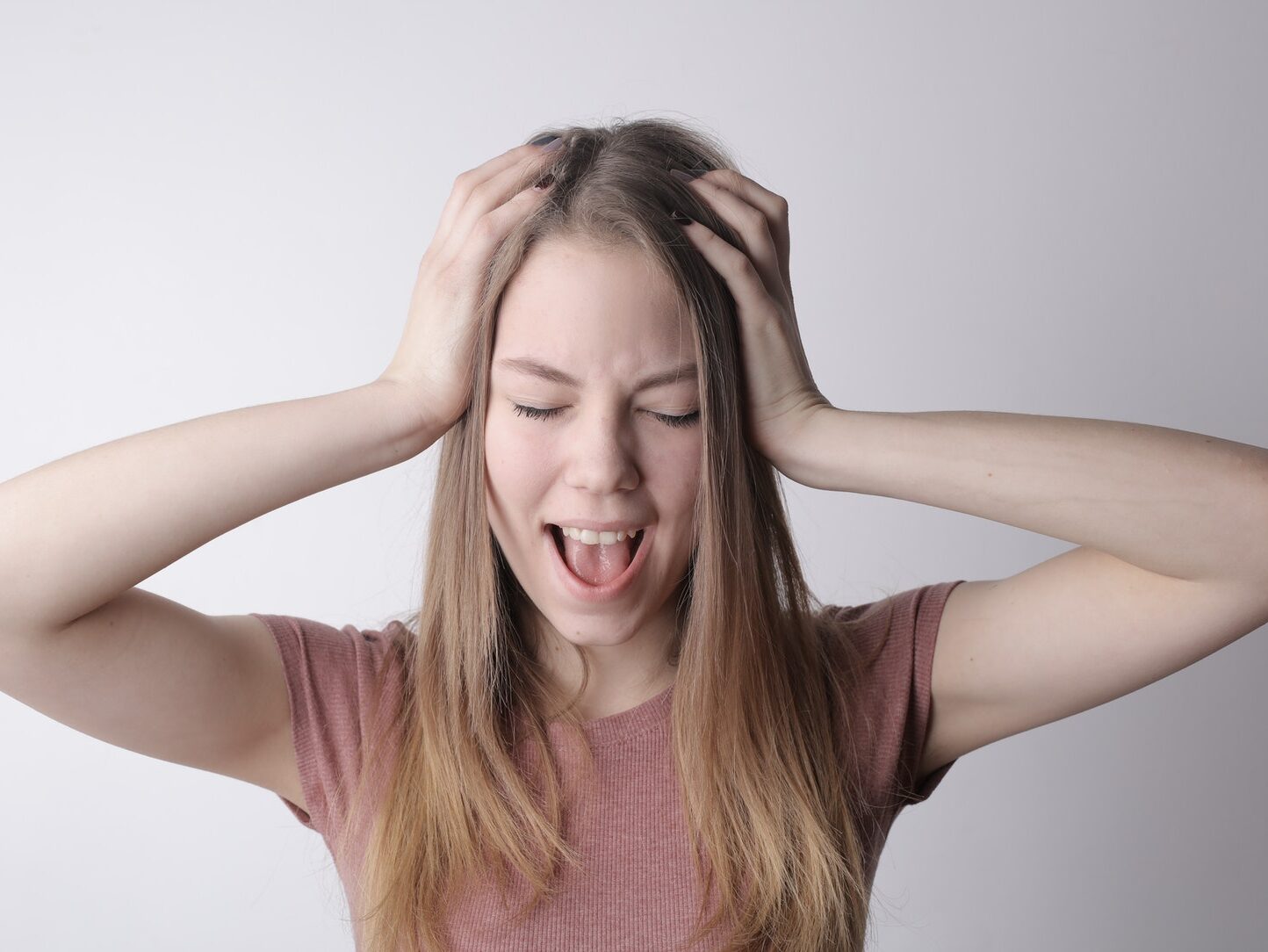 Angry crazy female in casual pink t shirt touching head while being unhappy and shouting loudly against gray wall background