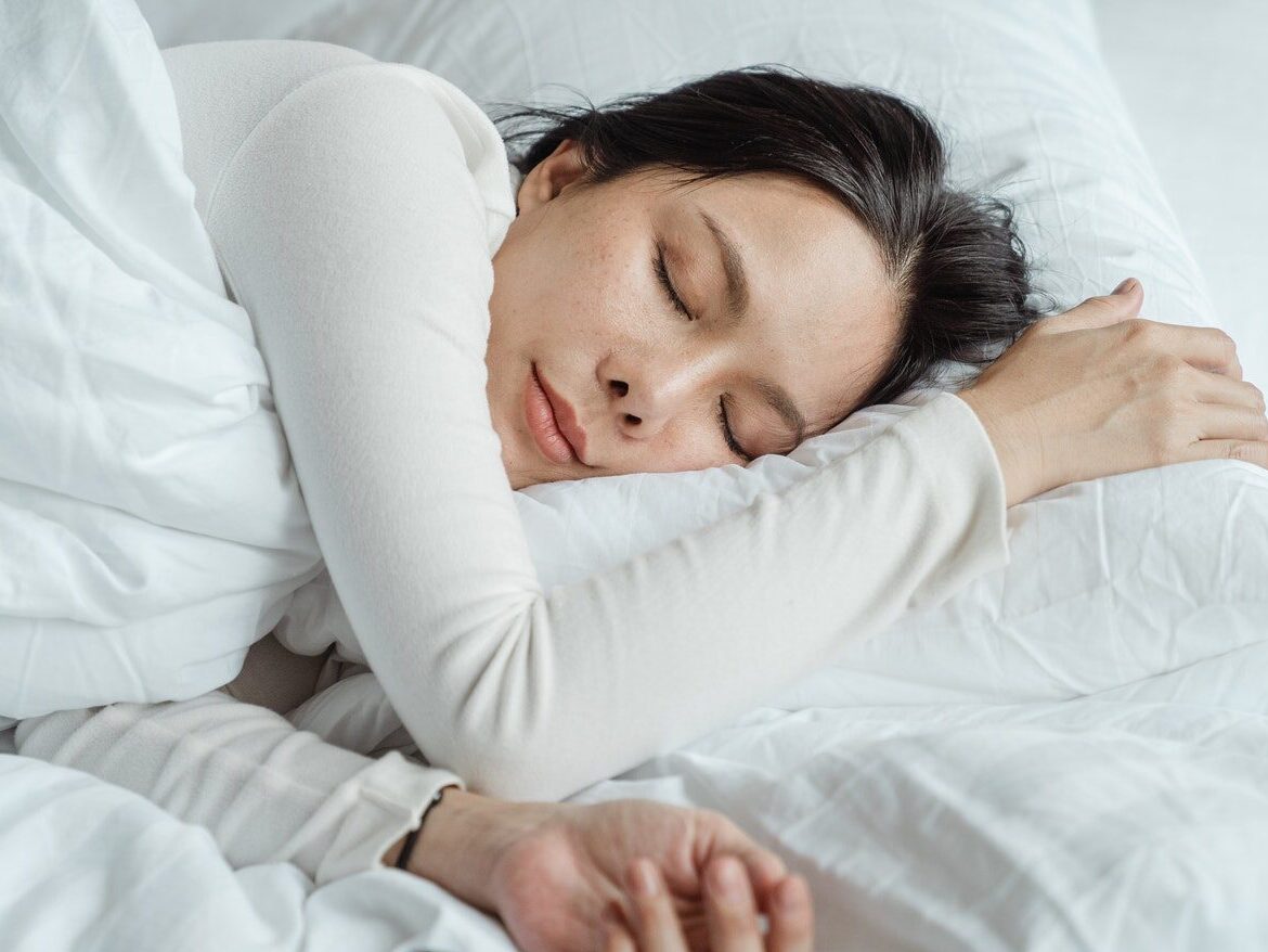 Calm Asian female wearing white pajama sleeping in comfortable bed with white sheets near modern mobile phone in morning