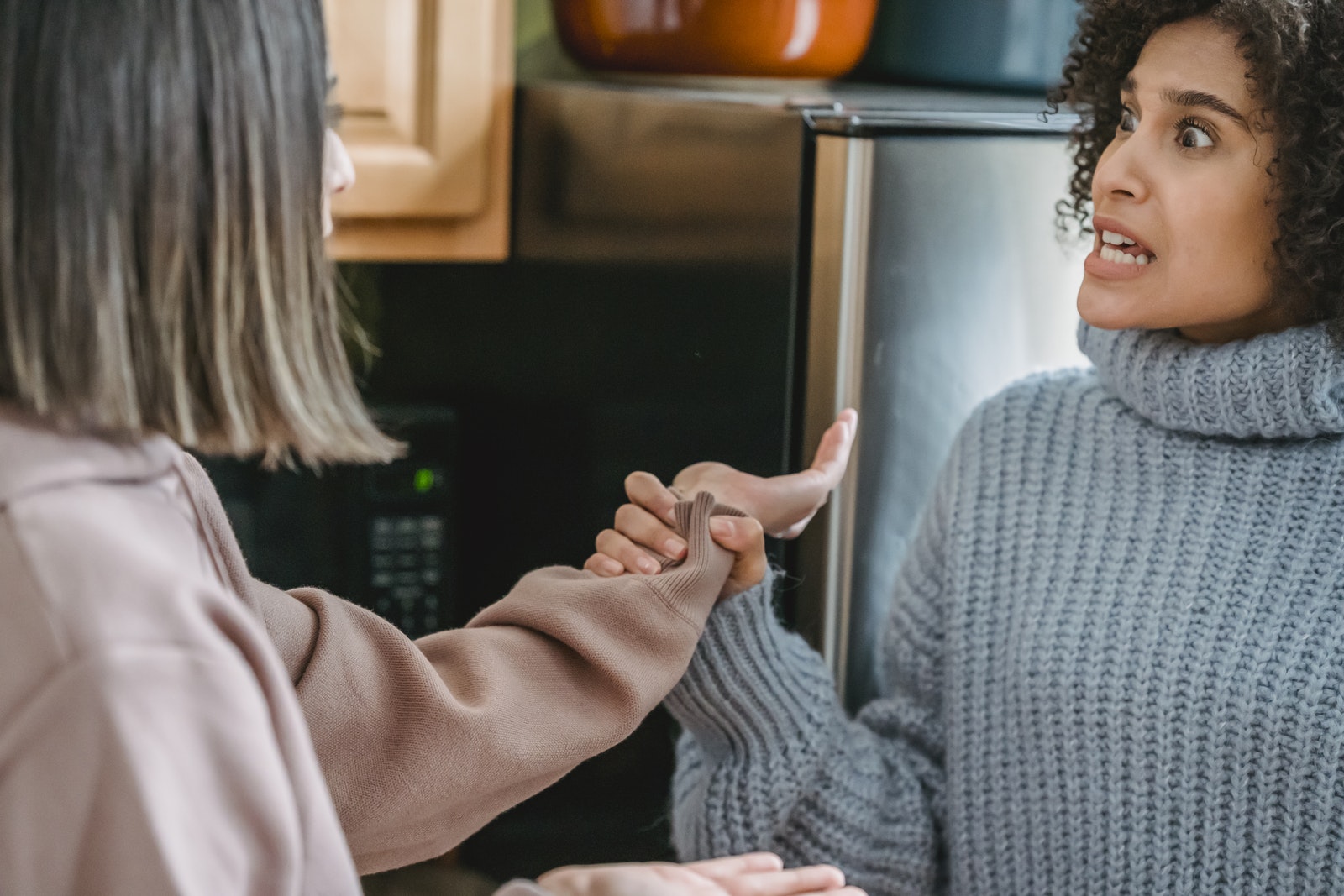 Angry African American female in casual wear shouting and grabbing hand of anonymous female while fighting and looking at each other in kitchen