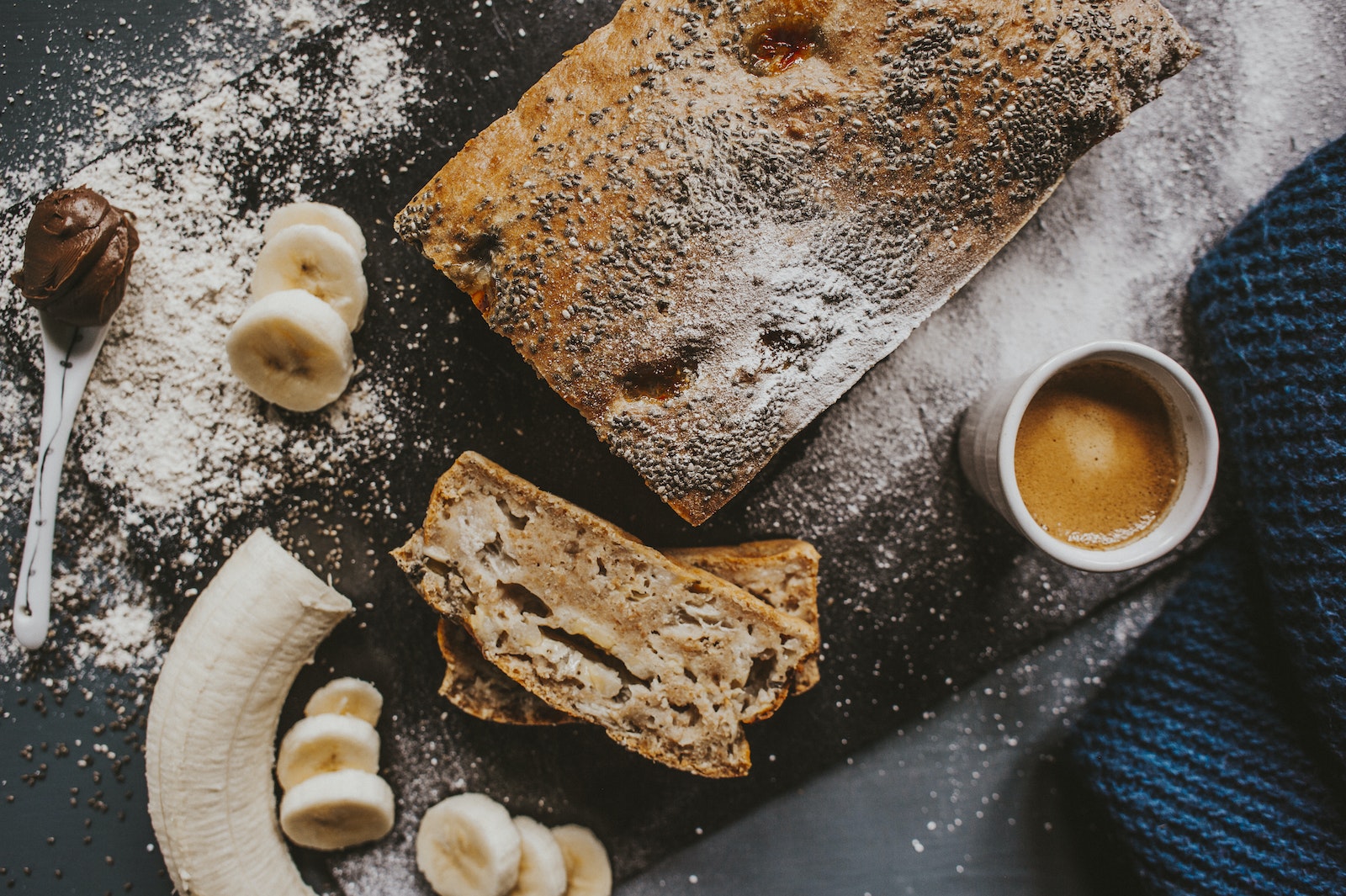 From above of appetizing cake topped with poppy seeds placed near cup of coffee and cut banana served on scattered with icing sugar chopping board on table