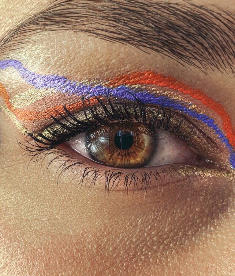 Closeup brown eye and eyebrow of anonymous female with bright purple and orange eyeliner with painted eyelashes looking at camera