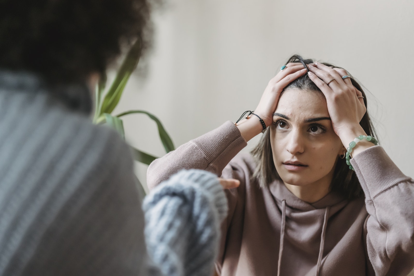 Worried female sitting near wall with hands on head and looking at faceless female while having conflict in light room