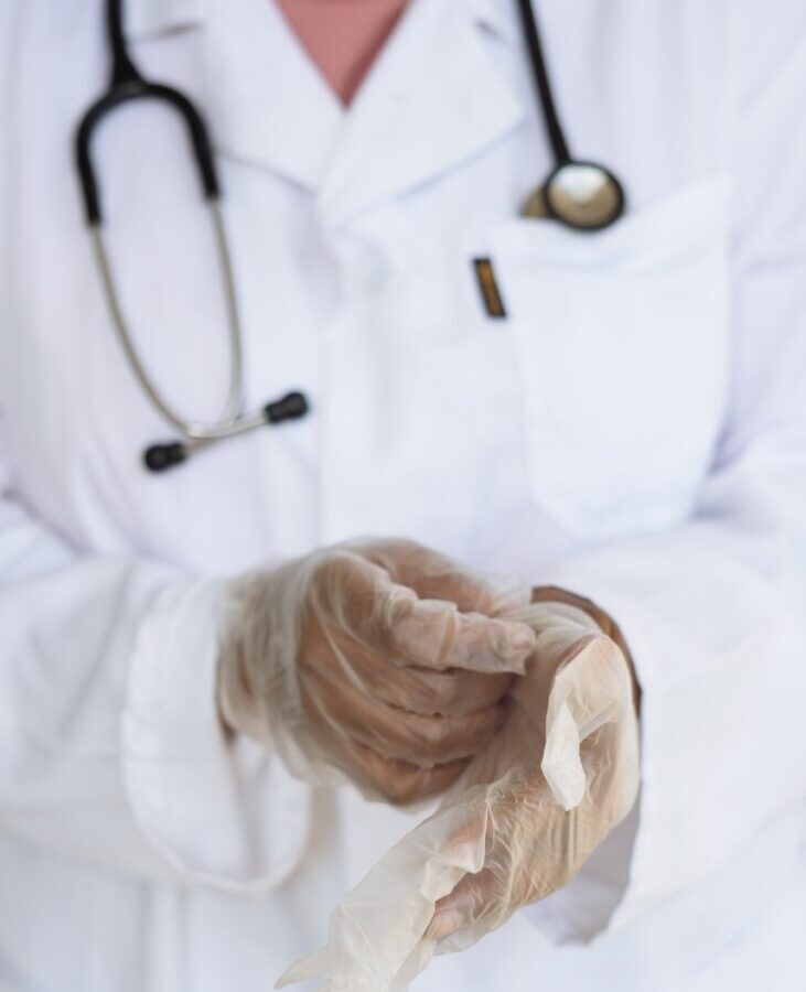 Faceless ethnic medical worker in lab coat and stethoscope taking of transparent gloves after approaching patients for examination in modern hospital