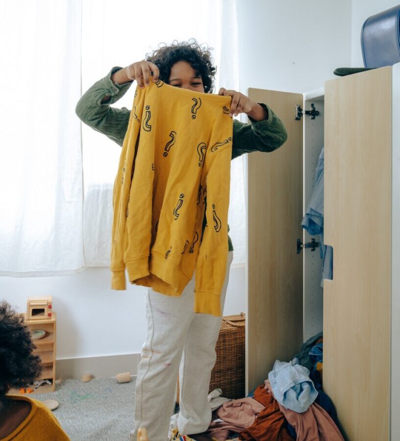 Unrecognizable African American teen girl in casual wear holding yellow sweater while sorting clothes in wardrobe in room