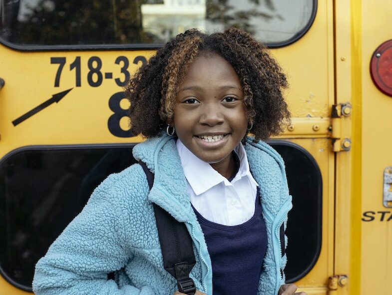 Cheerful African American girl with curly hair standing with backpack near yellow school bus and looking at camera with smile