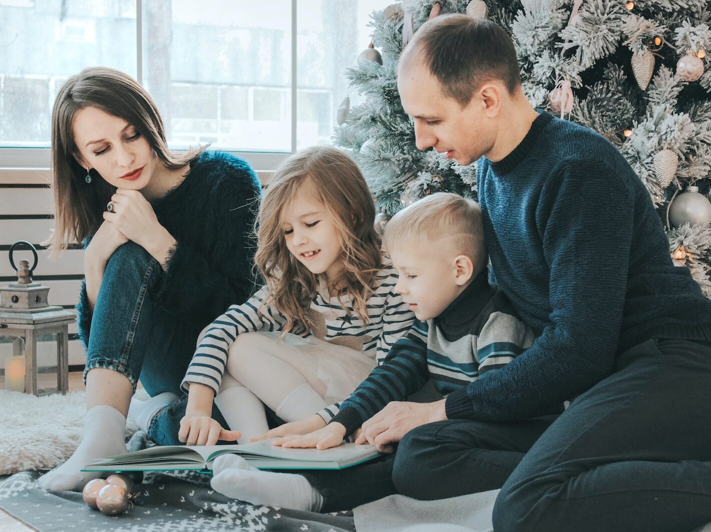 girl and boy reading book sitting between man and woman beside Christmas tree