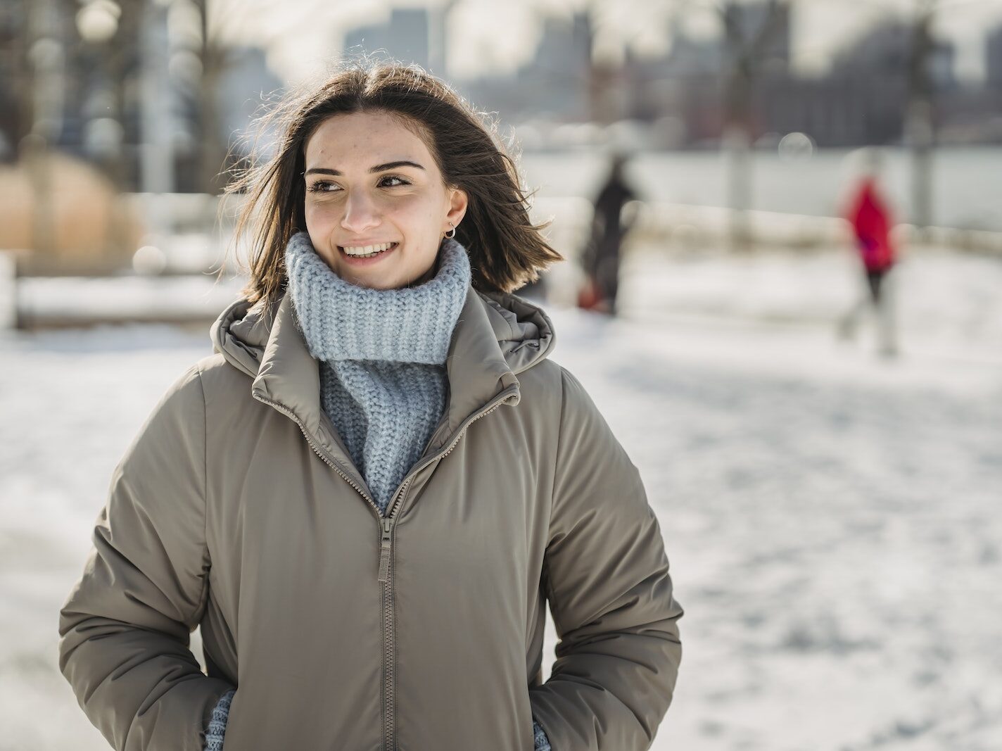Content young brunette in blue knitted sweater and warm jacket standing with hands in pockets in snowy city park on cold winter day