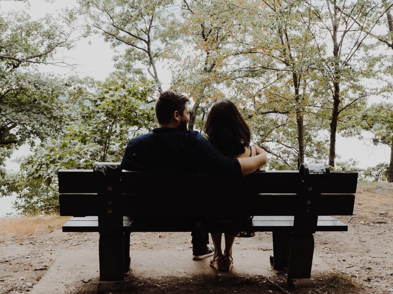 man looking to woman sitting on black wooden bench in front of tall trees during daytime