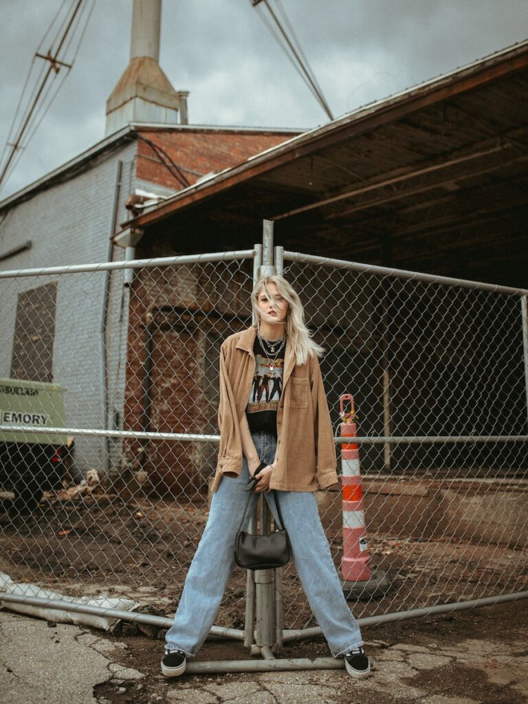 Young Blond Woman in Jeans Leaning against Grid Metal Fencing