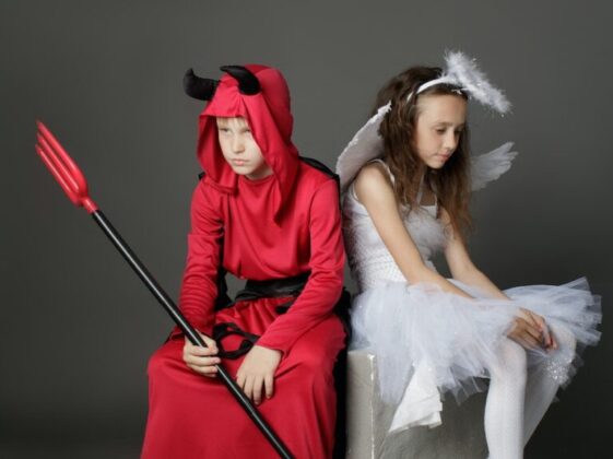 A Young Boy and Girl Wearing a Devil and Angel Costumes while Sitting on the Chair Back to Back