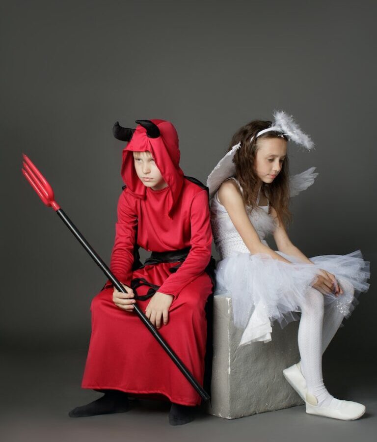 A Young Boy and Girl Wearing a Devil and Angel Costumes while Sitting on the Chair Back to Back