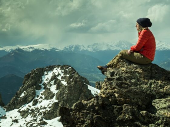 person sitting on rock across snow covered mountain under cloudy sky