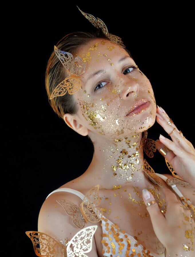 Young calm female model wearing white dress with sequins and golden butterflies touching skin covered with glittering golden foil while looking at camera during photo shoot in studio
