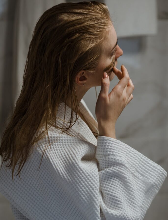 Young woman in bathrobe applying cosmetic mask on face