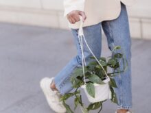 Crop anonymous female in stylish outfit and sneakers walking on sidewalk with hanging planter with green foliage on street in city