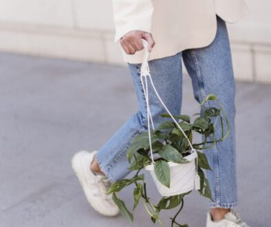 Crop anonymous female in stylish outfit and sneakers walking on sidewalk with hanging planter with green foliage on street in city